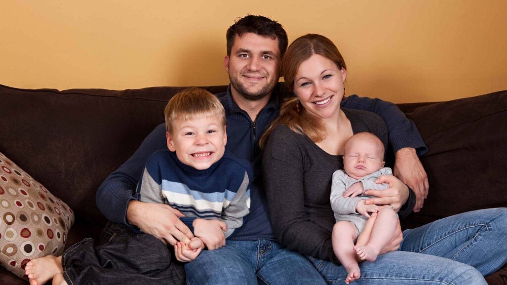 A smiling family sits on a blue couch. The father has dark hair and a partial beard and has one arm around a blond boy in a striped shirt. The other arm is around the woman, who has a long brown ponytail and is holding a sleeping infant in her arms.