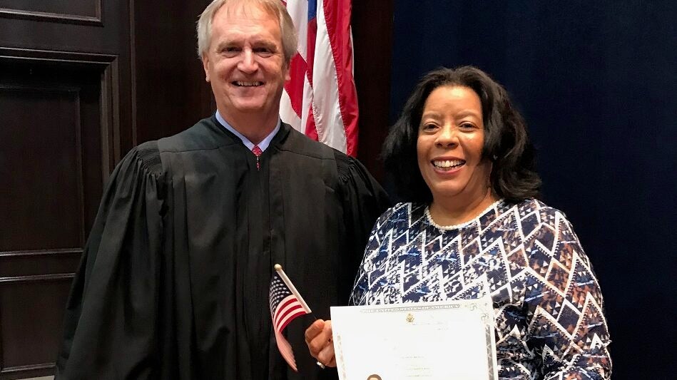 A judge in his robe stands with a woman in a sweater who holds a small American flag and a certificate of U.S. citizenship.