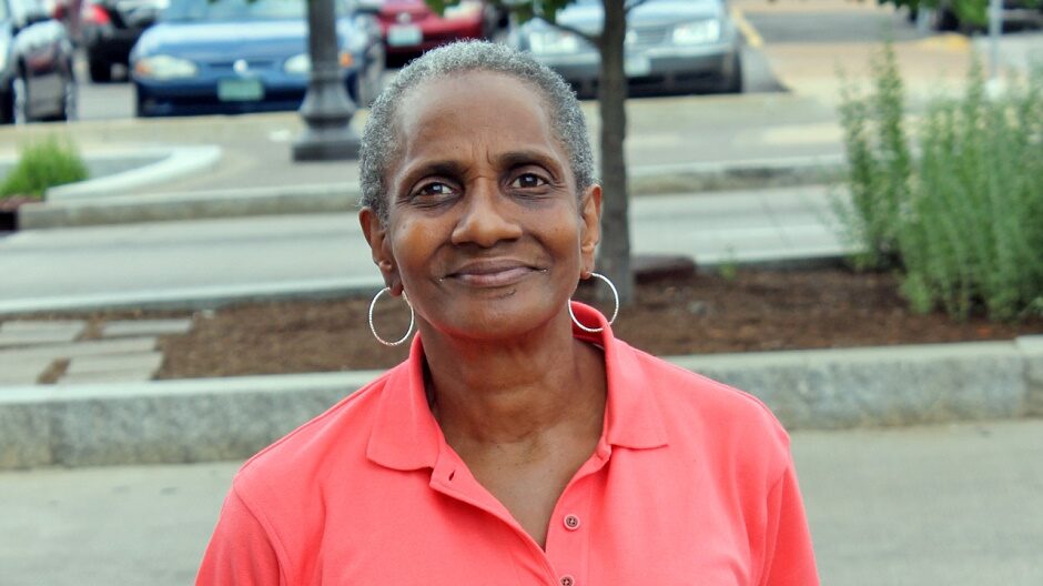 A woman with short cropped grey hair and silver hoop earrings looks directly at the camera with a half smile. She stands outside in a coral colored polo short.