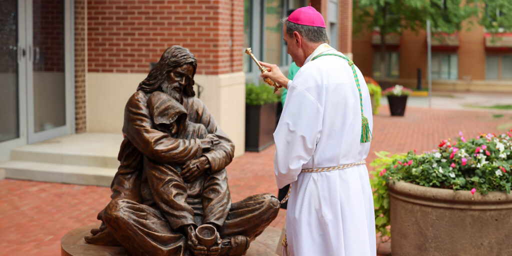 Bishop Michael F. Burbidge of the Diocese of Arlington blesses the sculpture "Hold It Together," by Christian sculptor Timothy Schmalz. The bronze sculpture depicts Jesus hugging a homeless man, who sits cross-legged on the ground, a hoodie obscuring his face, a cup in his hand for donations.
