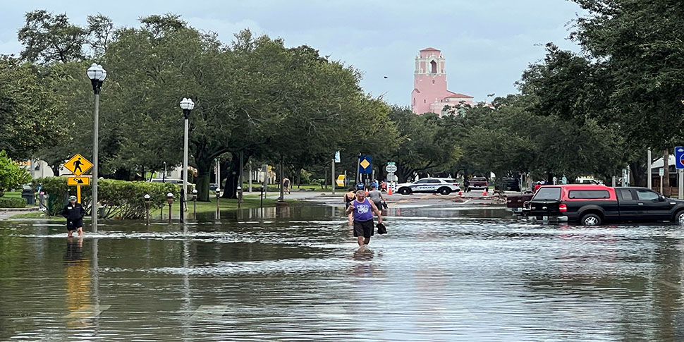 A man in a purple tank top wades through knee-high water after flooding caused by Hurricane Idalia. Several cars are in standing water halfway up their tires.
