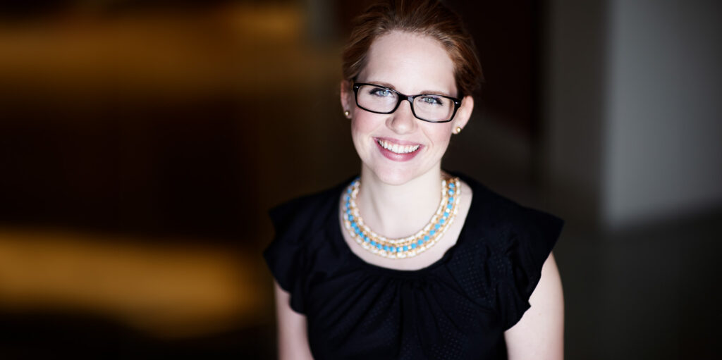 Photo of Sarah Clement, chief administrative officer at Catholic Charities of Acadiana. She has red hair, a broad smile, black rim glasses, a black top and a turquoise necklace.