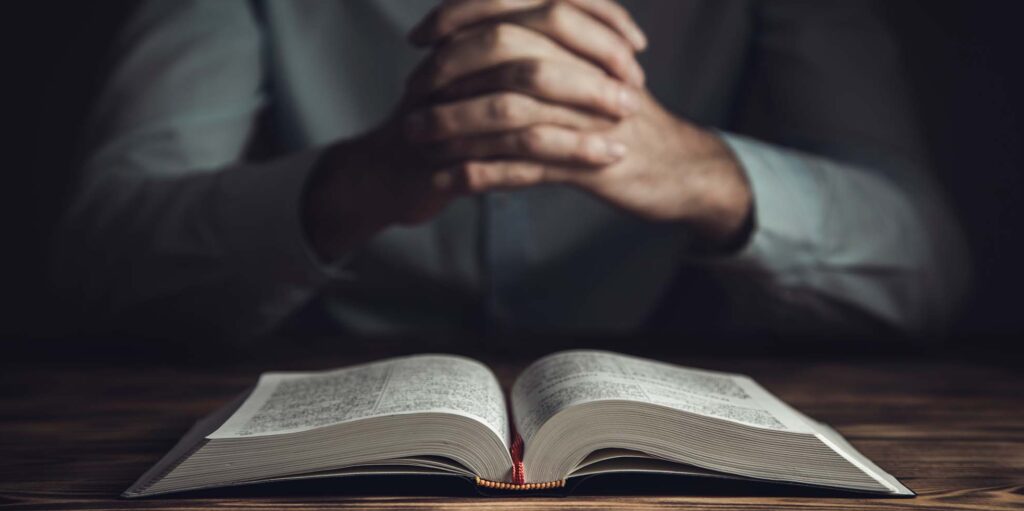 Close-up of a man's hands clasped in prayer. A Bible is open on the table before him.
