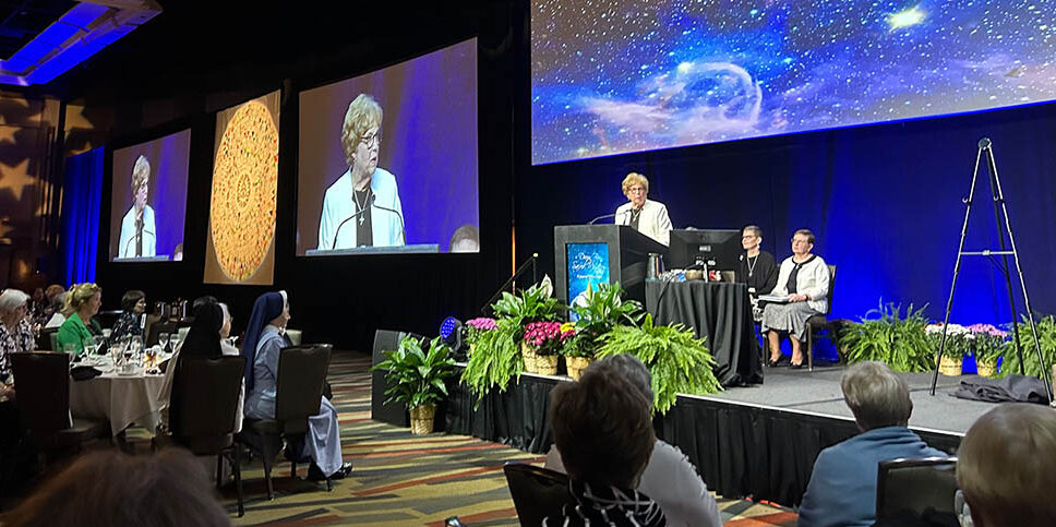 A photo from a Catholic Charities USA annual Gathering. Then-president and CEO Sr. Donna Markham is on the stage addressing the crowd. There are two woman sitting behind her and plants on the stage. The audience is seated at round tables before her.