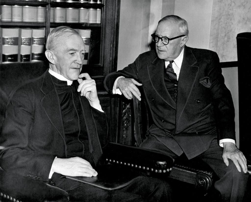 An historic photo of Fr. John J. O'Grady with Senator Robert Wagner in 1945. They are seated in leather armchairs in front of a bookcase filled with law books. The senator is looking at Fr. O'Grady, who sits pensively with his hand to his jaw.