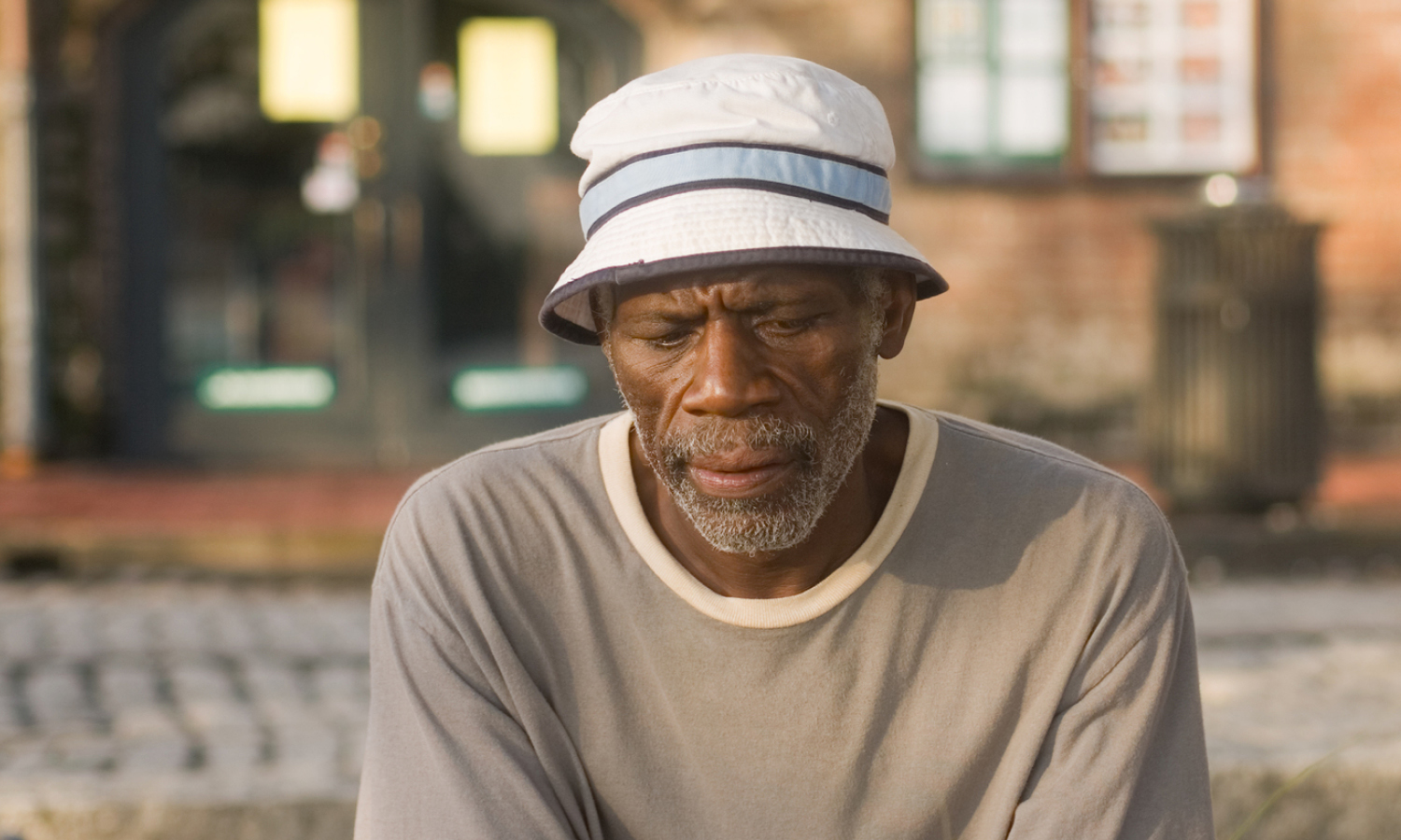 An older man with a hat sits outside, looking downcast. 