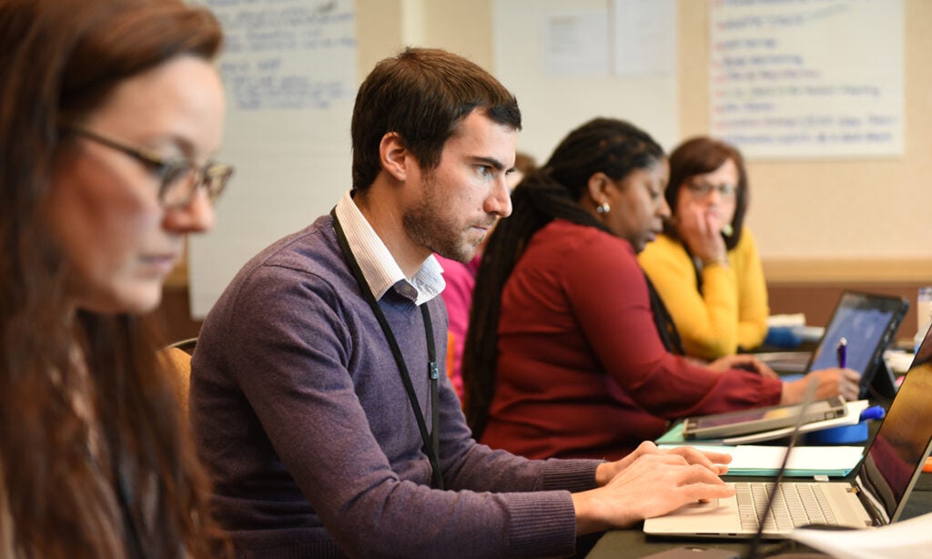 A young man with a purple sweater and a partial beard works on a laptop computer. Two women sit at the table to his left and another sits to his right. They appear to be at a conference.