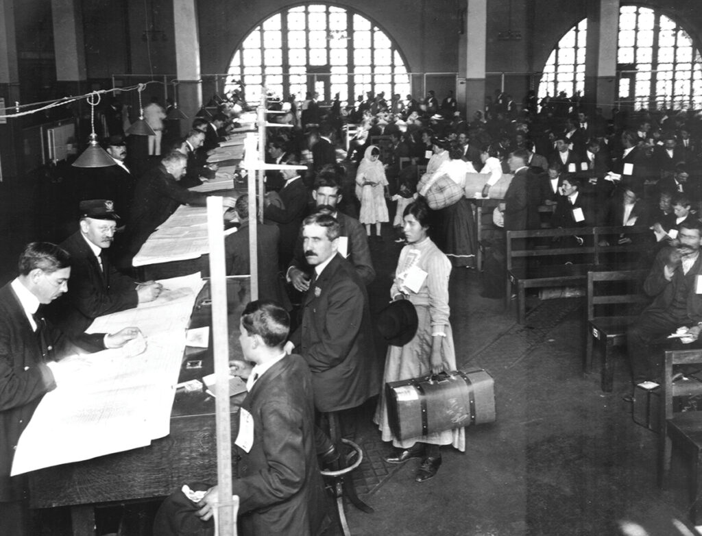 An historic photo of people lined up to be processed at Ellis Island. The men wear suit jackets and mustaches, and a woman stand in the foreground, carrying a suitcase with her dark hair piled on top of her head.