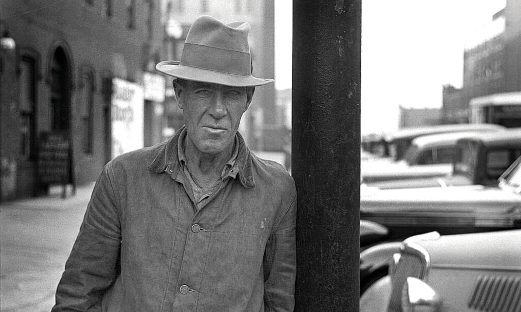 A Depression-era man in a fedora leans against a telephone pole in this black and white photos. 