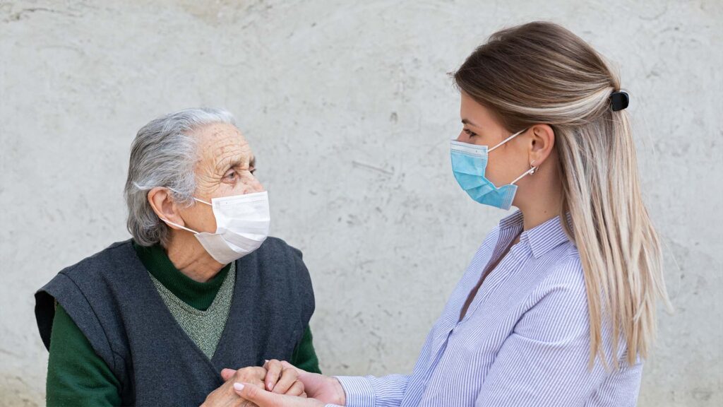 An old woman wearing a covid mask holds hands with a young woman, also wearing a mask. They are looking at each other. The younger woman might be a medical professional.