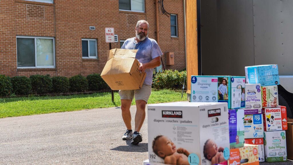 As part of a Hurricane Ida recovery effort, a bearded man in a t-shirt and shorts carries a box of diapers toward the back of a truck, where other boxes of diapers are piled.
