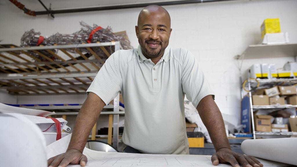 A smiling man stands with both hands on a table before him, on top of architectural plans. Behind him are shelves with boxes and construction supplies.