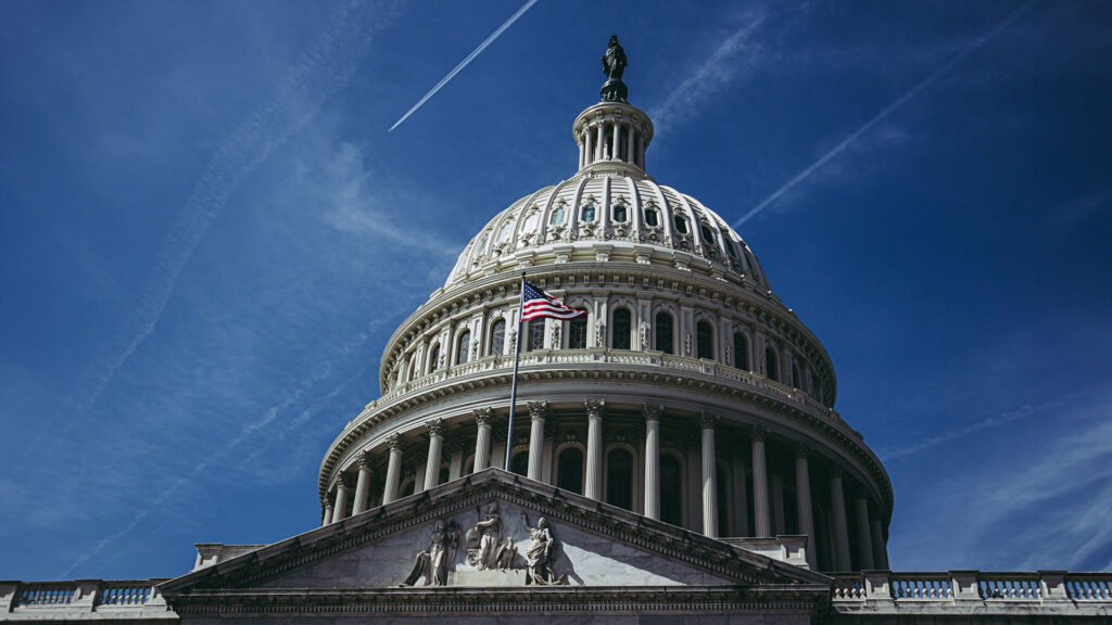 A close-up of the U.S. Capitol Dome with the U.S. flag flying and streaks of airplane trails in the sky above.