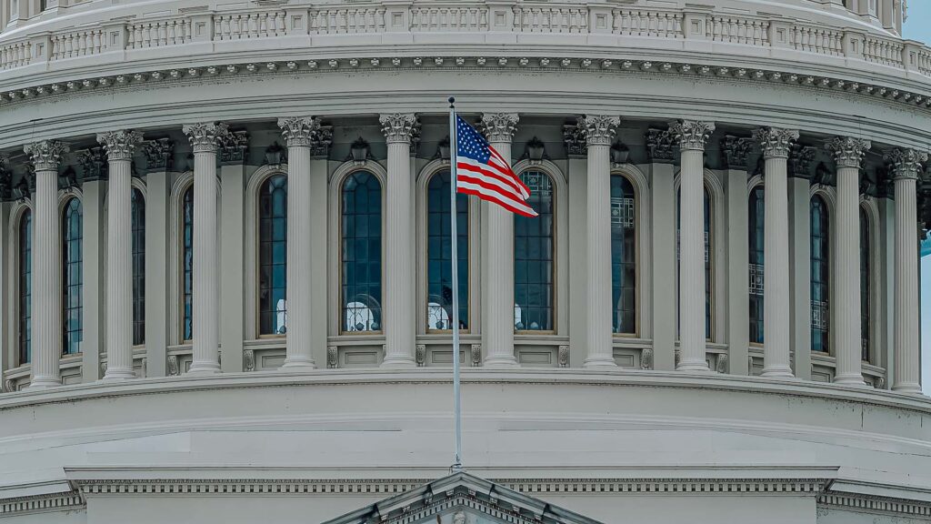 A close up shot of rotunda of the US Capitol building, with an American flag waving in the front.