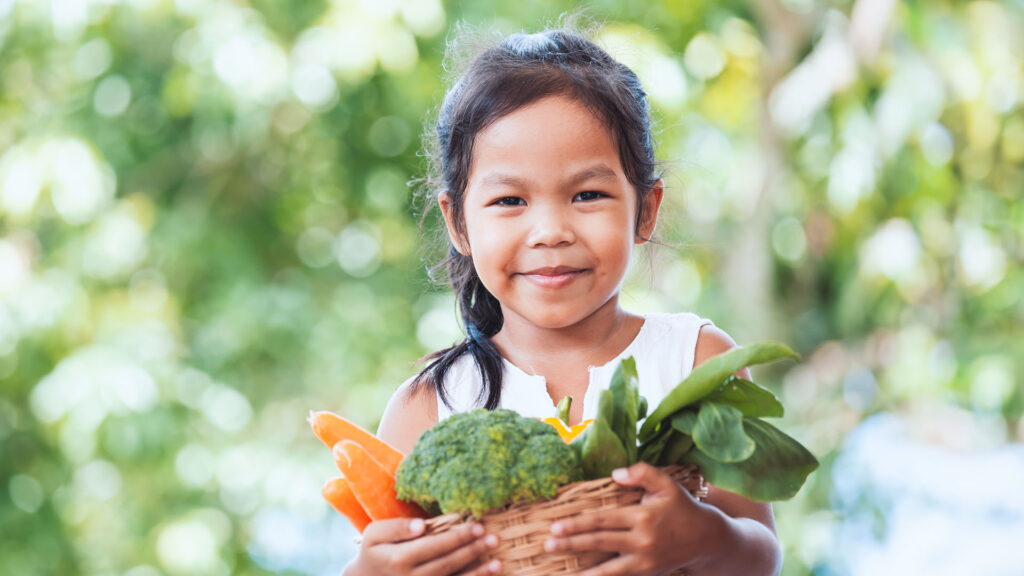 A smiling little girl with dark hair holds a basket with fresh produce -- carrots, broccoli and greens -- spilling out over the top.