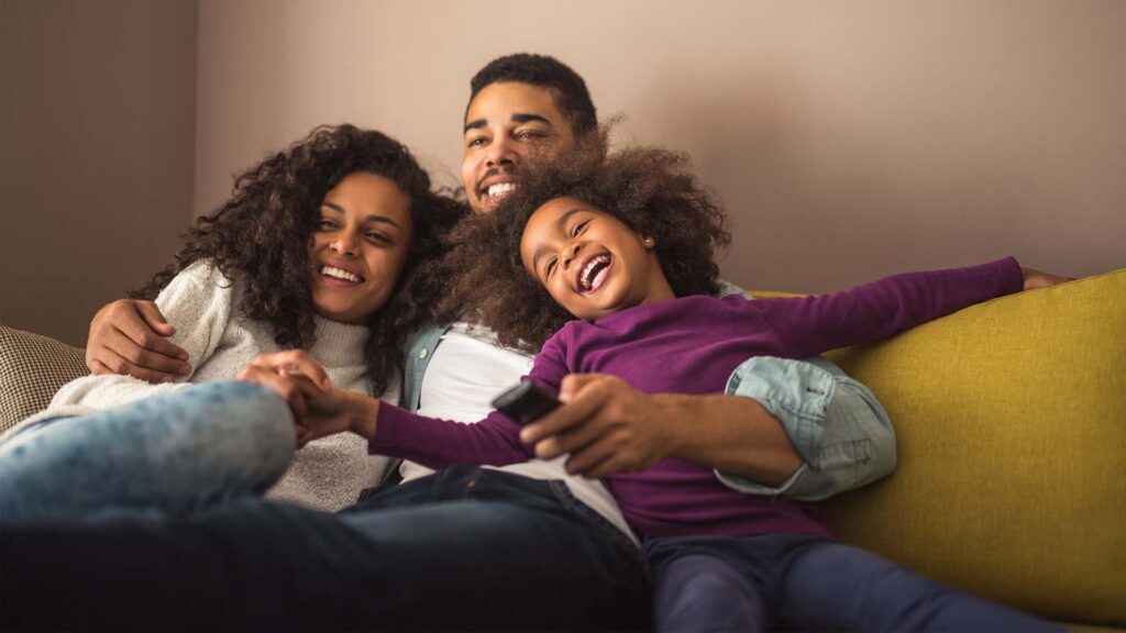 A happy family sits on the couch together. The man has one arm around a smiling woman, who leans into him, and the other around a little girl in a purple shirt, who leans against him. The woman and girl are holding hands, and the man has a remote control in one hand.