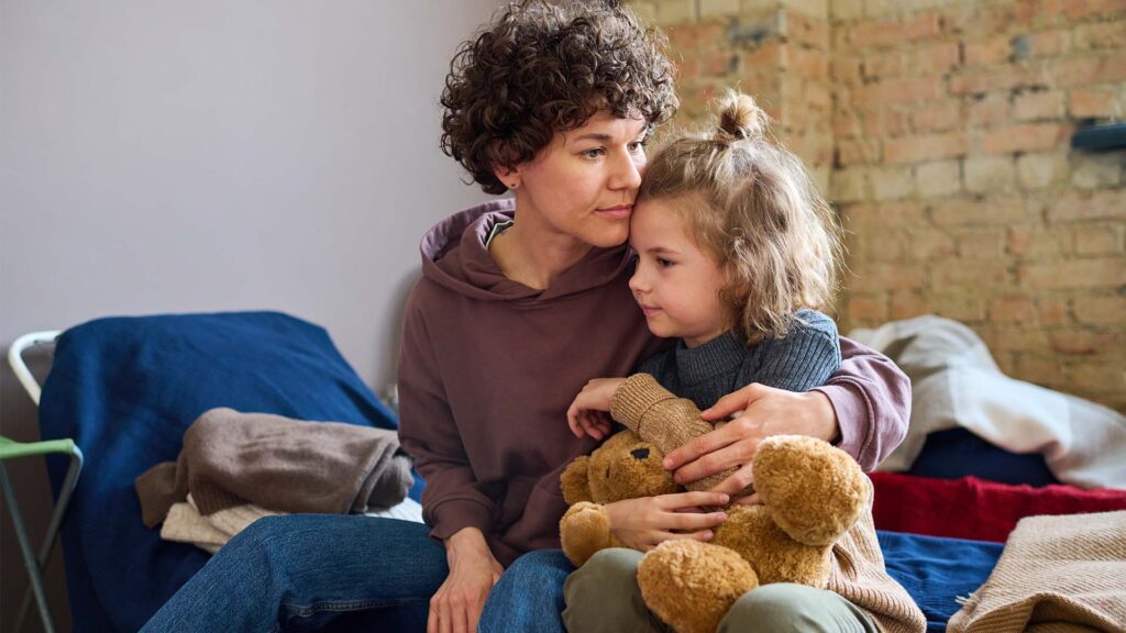 A woman hugs her little girl while sitting on a cot at a Catholic Charities shelter. The woman looks pensive, and the girl is holding a teddy bear.