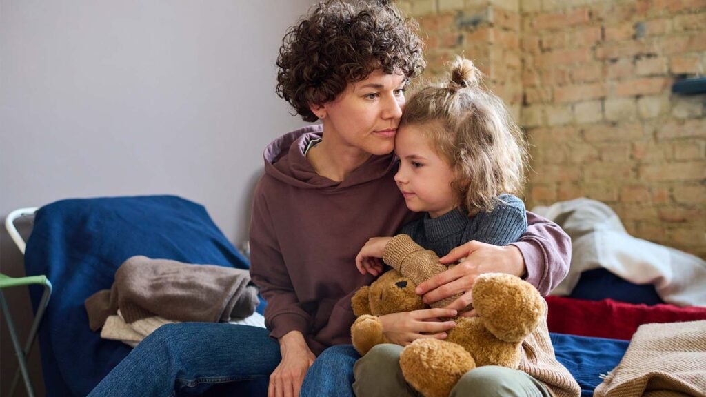 A woman hugs her little girl while sitting on a cot at a Catholic Charities shelter. The woman looks pensive, and the girl is holding a teddy bear.