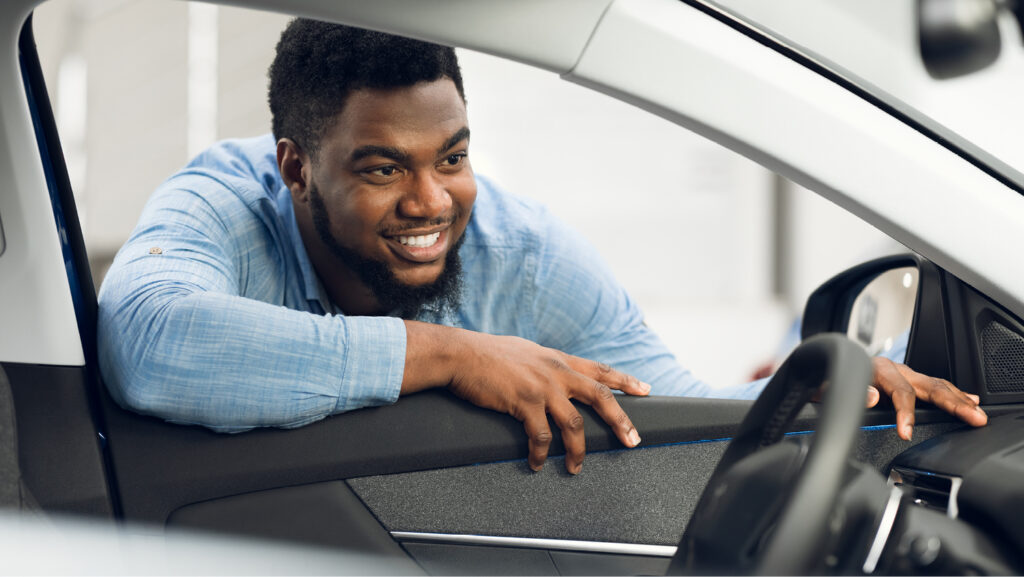 A smiling young man with a beard and a blue denim shirt leans in the driver's side window of an empty car. 