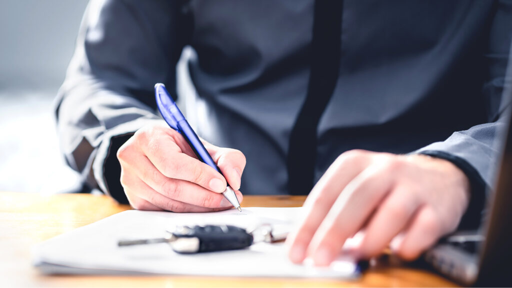A person signs a document with a blue pen. A car key rests on the stack of papers.