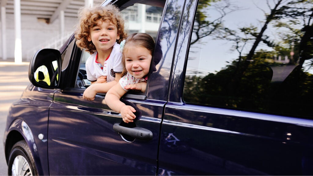 A little boy and girl lean out the driver's side window of a blue parked car. They're smiling in a donated vehicle.