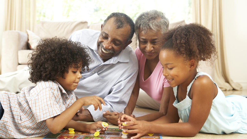 Grandparents and two grandchildren, a boy and a girl, lie on the living room floor, playing a board game. They're all smiling and happy. 