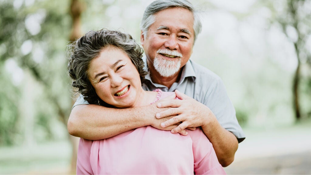 A smiling older couple stand together. He has a goatee and his arms around the woman, who wears a pink top. 