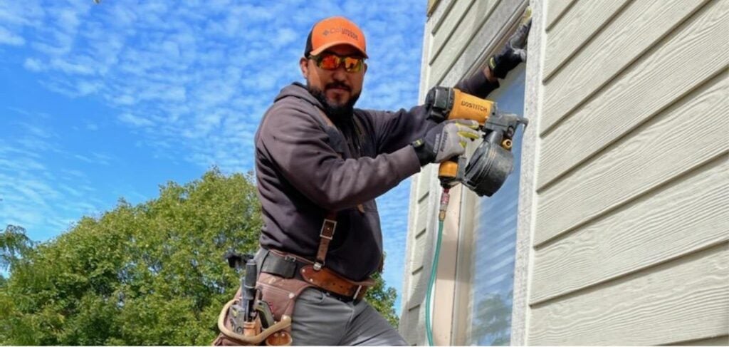 A man in an orange cap, mirrored sunglasses, blue hoodie and a tool belt holds a large tool against a window frame. He is working construction.