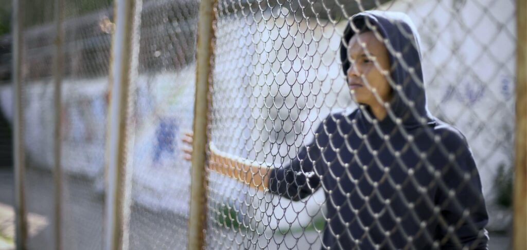 A young man in a dark blue hoodie stands on one side of a chain link fence, looking through it.