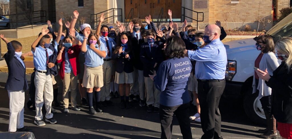 A group of Catholic elementary school students stand outside their school, waving and clapping.