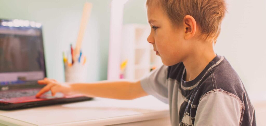 A little boy in a t-shirt sits at a desk. His right hand stretches out to the keyboard of a laptop.