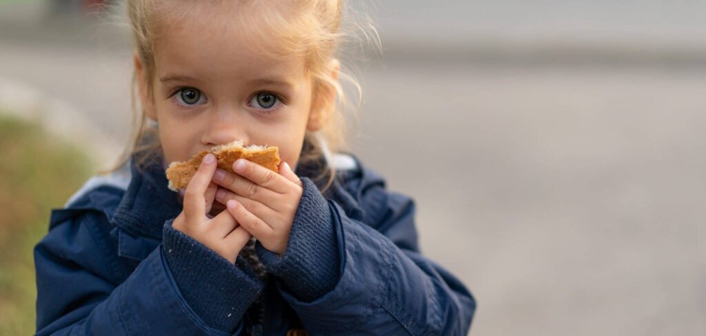A little girl with large blue eyes and blonde hair holds a piece of bread to her mouth. She's wearing a dark blue coat.