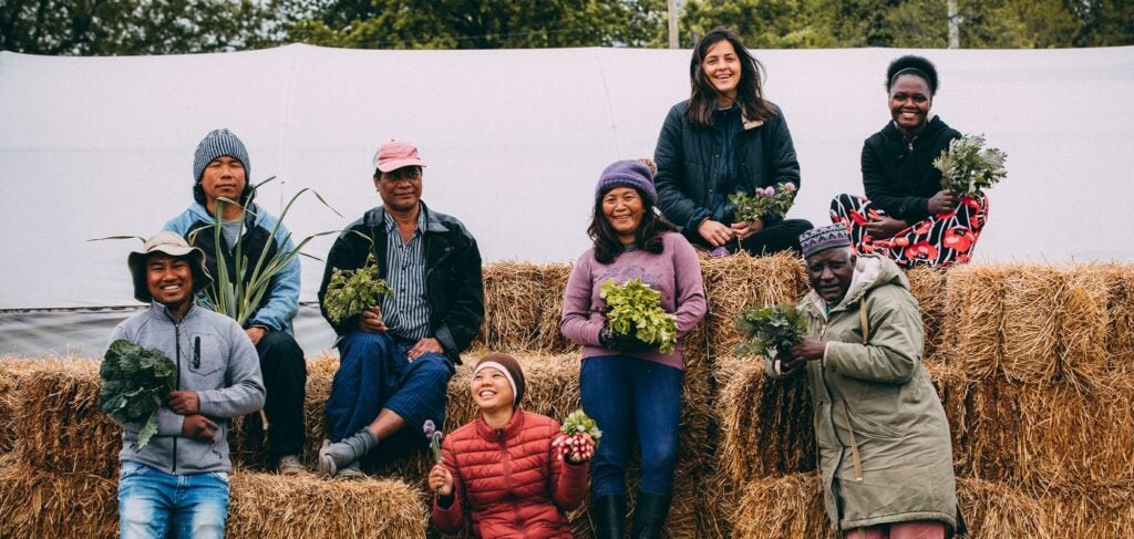 A group of eight participants in the New Roots for Refugees garden program of Catholic Charities Northeast Kansas. They sit on bales of hay holding various produce -- lettuce, carrots, herbs, etc.