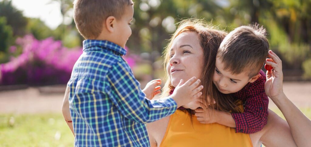 A woman in a yellow top plays with two little boys. One, in a red checked shirt, leans over her shoulder with his arms around her neck. The other, in a blue check shirt, stands in front of her with his hands reaching out toward her face.