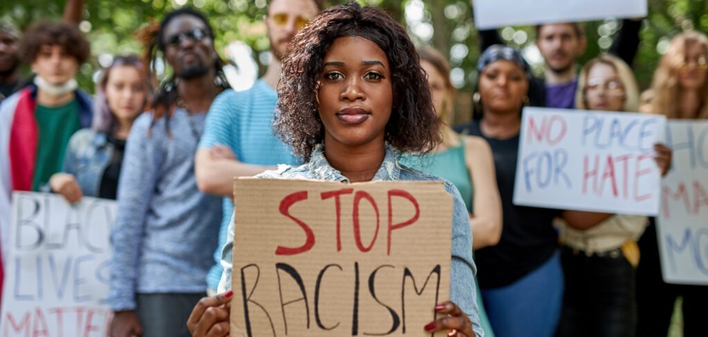 A young African-American woman stands before a crowd of people. She holds a sign that reads "stop racism," while those in the background hold signs saying "Black Lives Matter" and "No place for hate."
