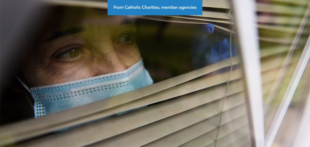 An older woman wearing a covid mask peers out through window blinds.