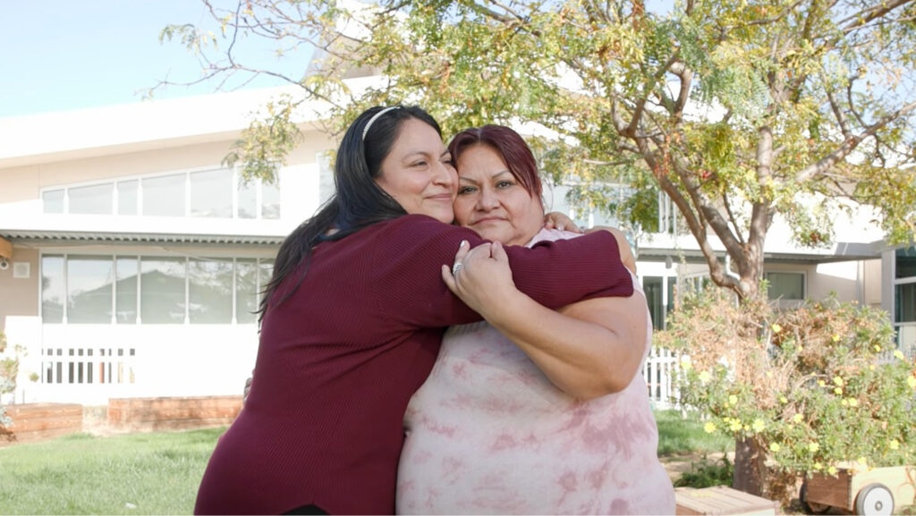 Photo of two women hugging each other in front of a church providing Foundational Services to the community.