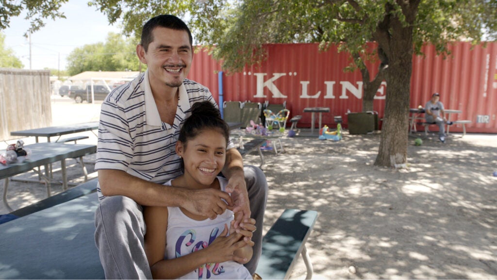 A smiling man and woman sit outside at a picnic table. He has his arms over her shoulders. They're in a sunny courtyard with a tree and children's play things scattered behind them. 