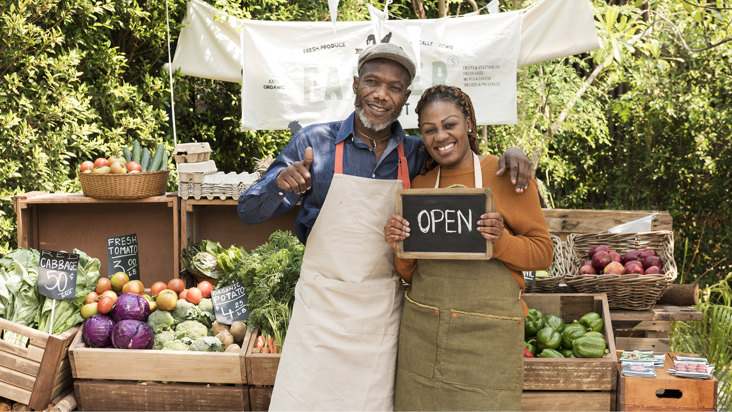 A happy couple in aprons stands in front of a produce stand. He has his arm over her shoulder and is giving a thumbs up while she holds a small chalkboard reading "Open." 