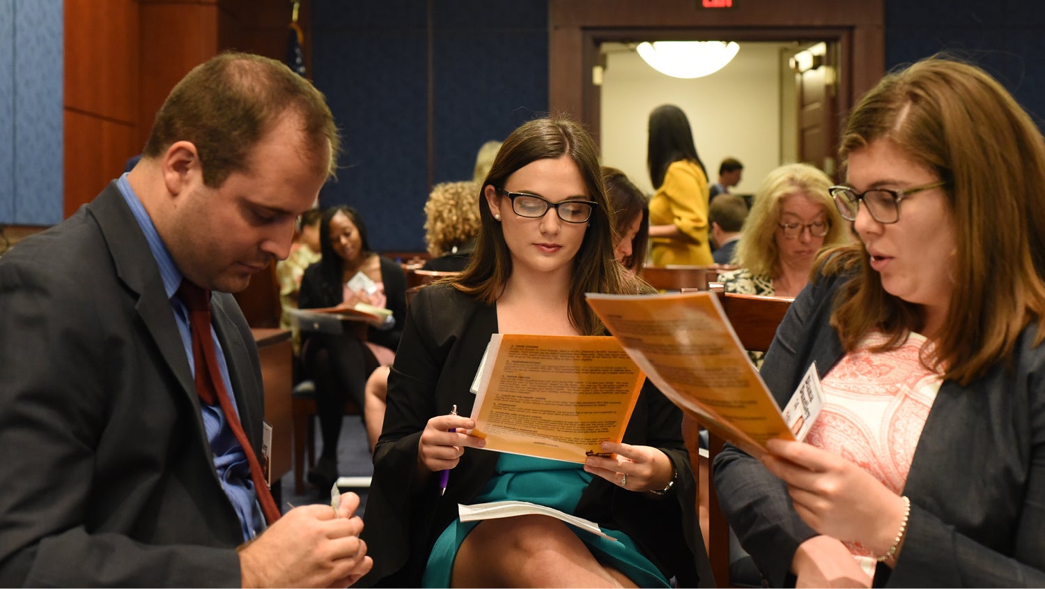 People wearing conference badges sit in a meeting room, reviewing paperwork. 