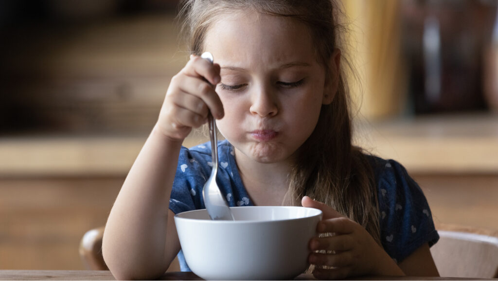 A little girl in a blue top with white heart holds a spoon and is about to take another bite of soup. 