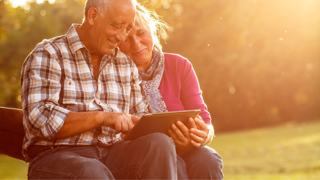 An older couple sit outside, looking at something on a computer tablet. She leans her head against his shoulder. The sun is shining on them.