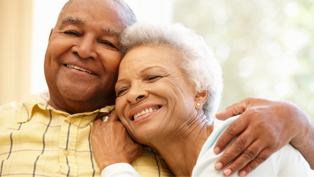 An older couple smile and look happy. Their heads are pressed together; she rests her hand on his chest and he has his arm around her shoulder.