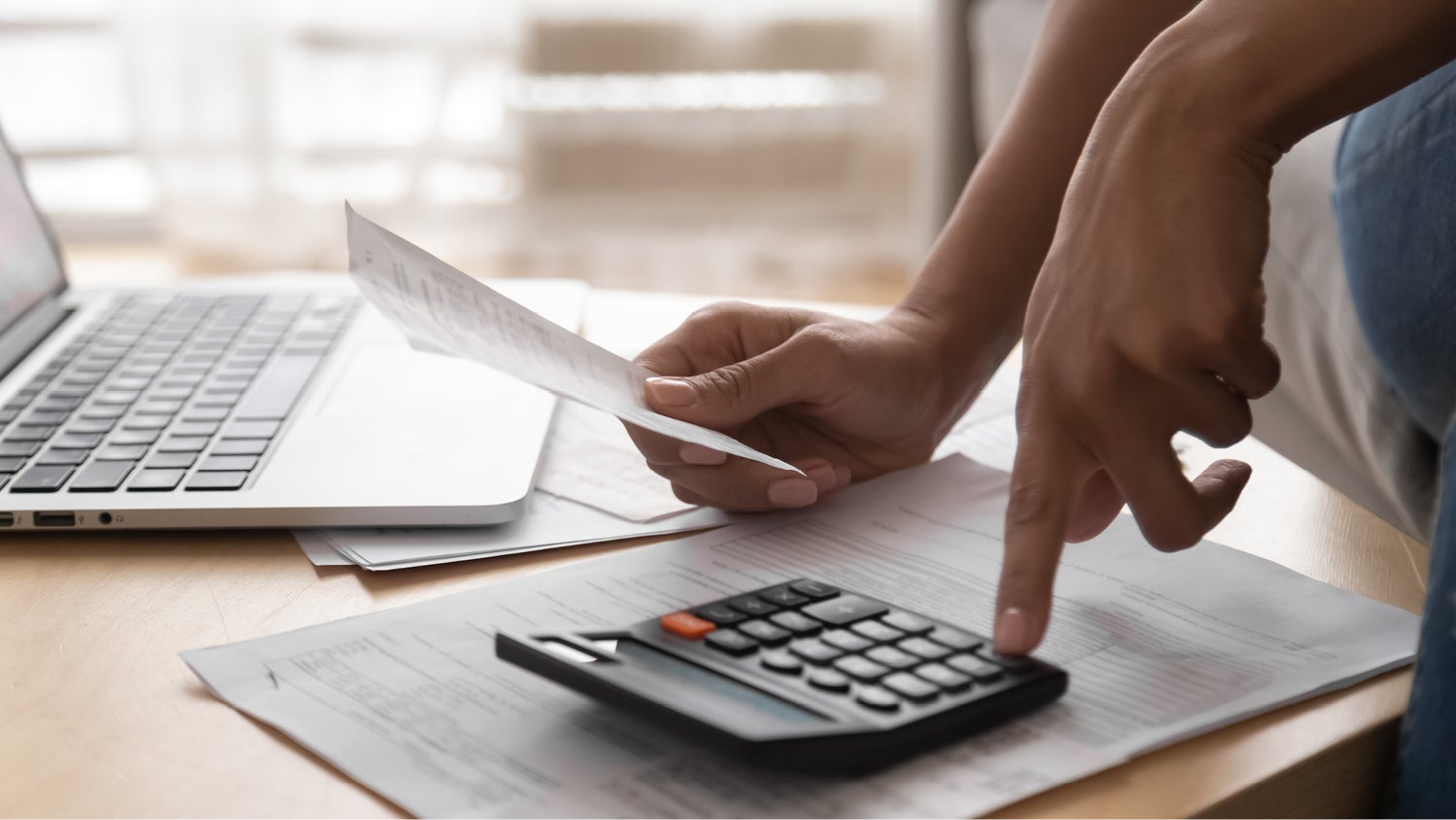 A person sits with a laptop on a coffee table, holding a receipt and working on a calculator.