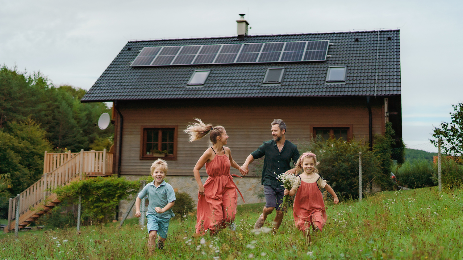A family of four run in a green field in front of a wooden house with solar panels on the roof. The parents hold hands and the boy and girl run ahead of them, the girl with flowers in her hand. 