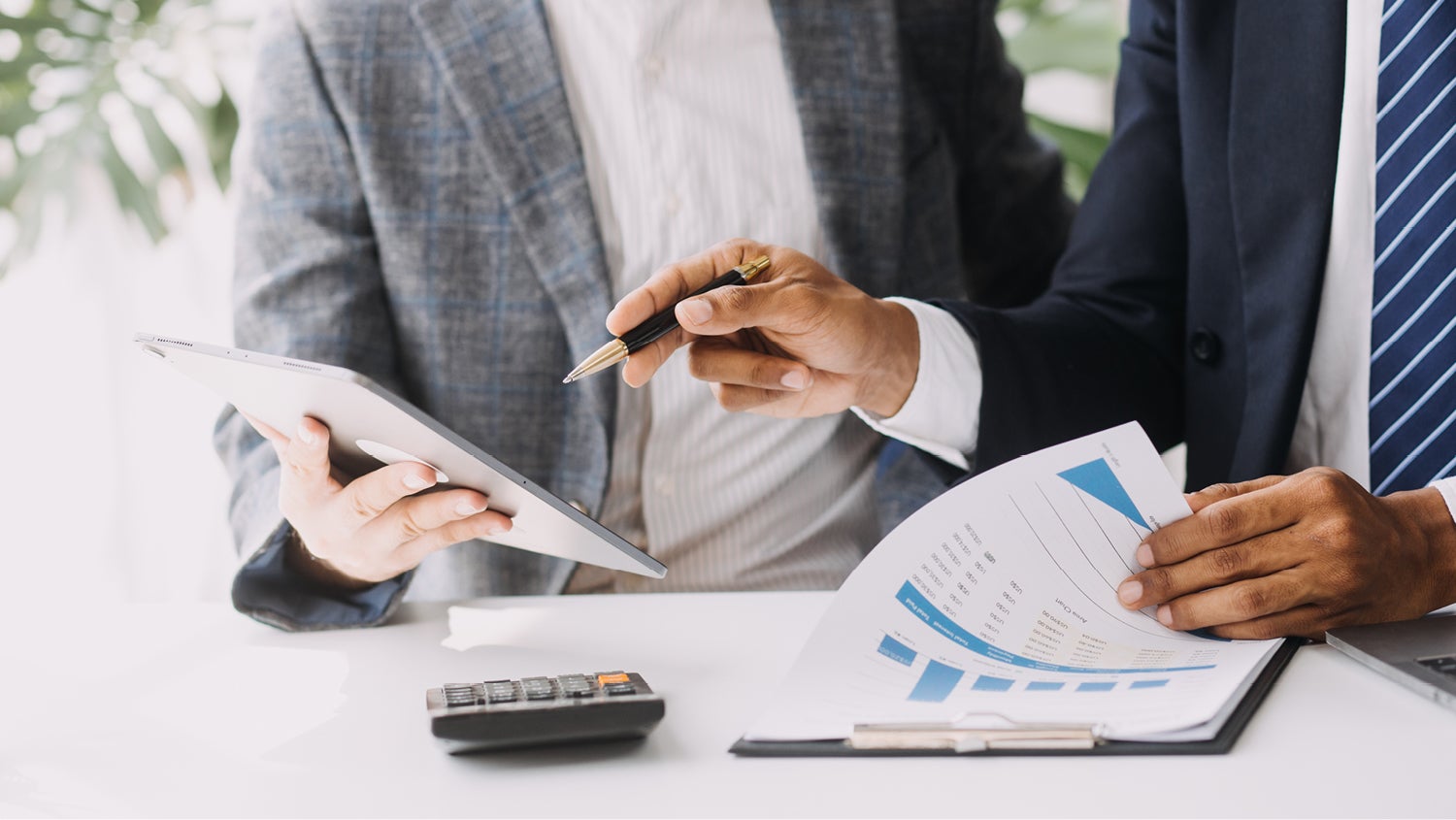 A closeup of the hands of two people going over financial paperwork. One holds a tablet computer, the other holds a pen and flips through paperwork. 