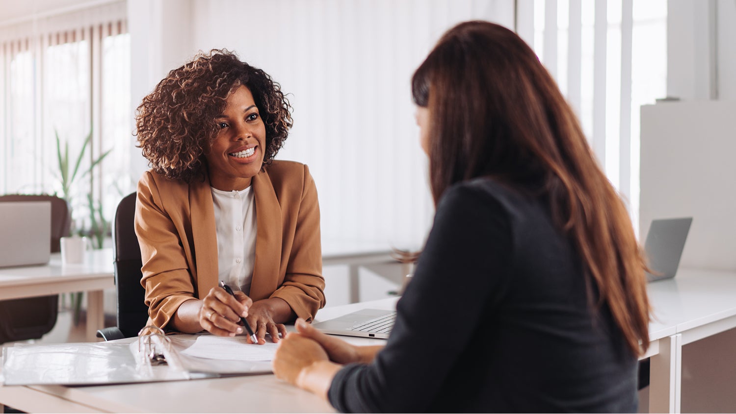 Two young women meet in a bank setting to go over paperwork. The employee is smiling at the other woman. 