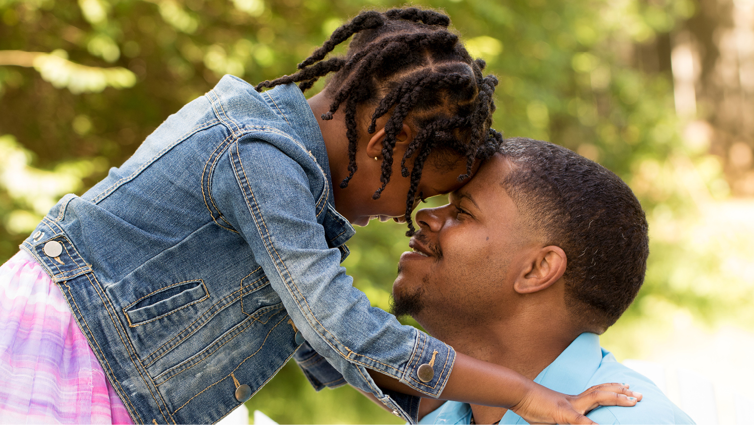 A little girl with braids in her hair wearing a jean jacket puts her forehead against her father's forehead. They are smiling at each other. 