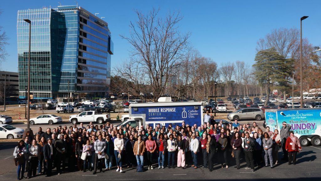 A group shot of the 225+ participants from more than 60 Catholic Charities agencies, nonprofits and government partners that took part in the 2023 Applied Institute for Disaster Excellence (AIDE) in Raleigh, North Carolina. They're standing in a parking lot. Behind them is the CCUSA Mobile Response Center vehicle.