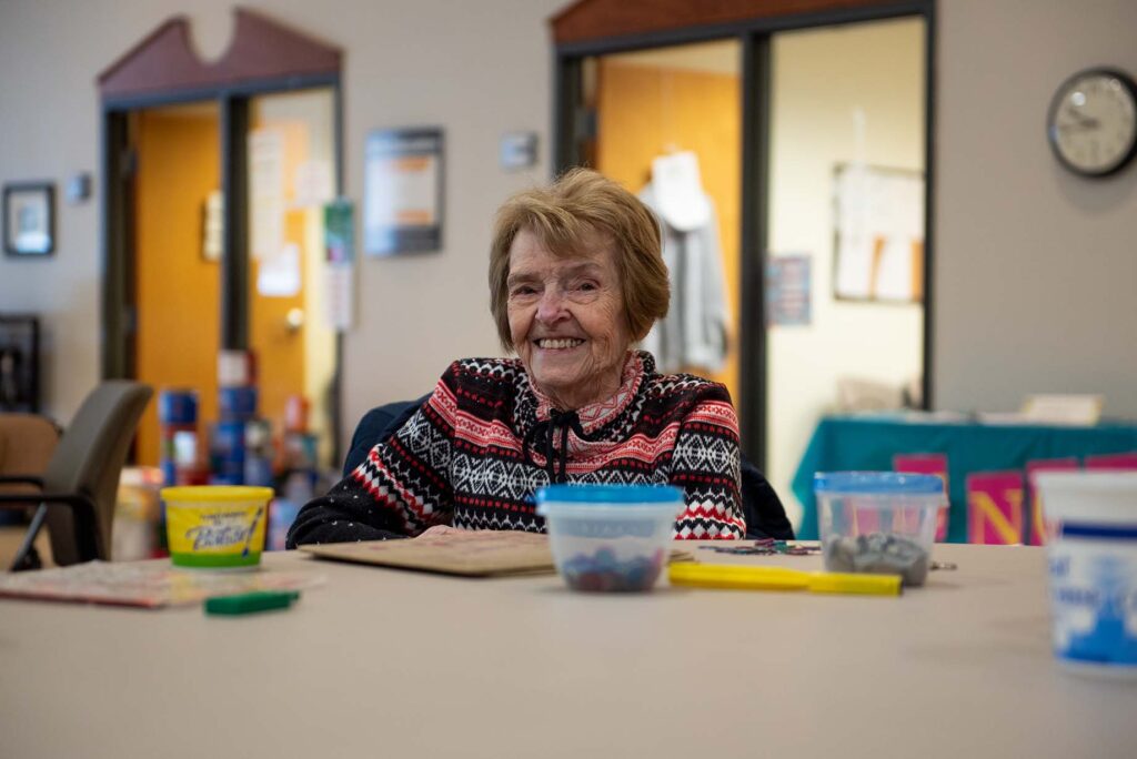 A smiling older woman in a patterned sweater sits at an activity table in a Catholic Charities facility.